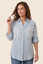 Woman wearing a light blue denim shirt with floral embroidery on a beige background
