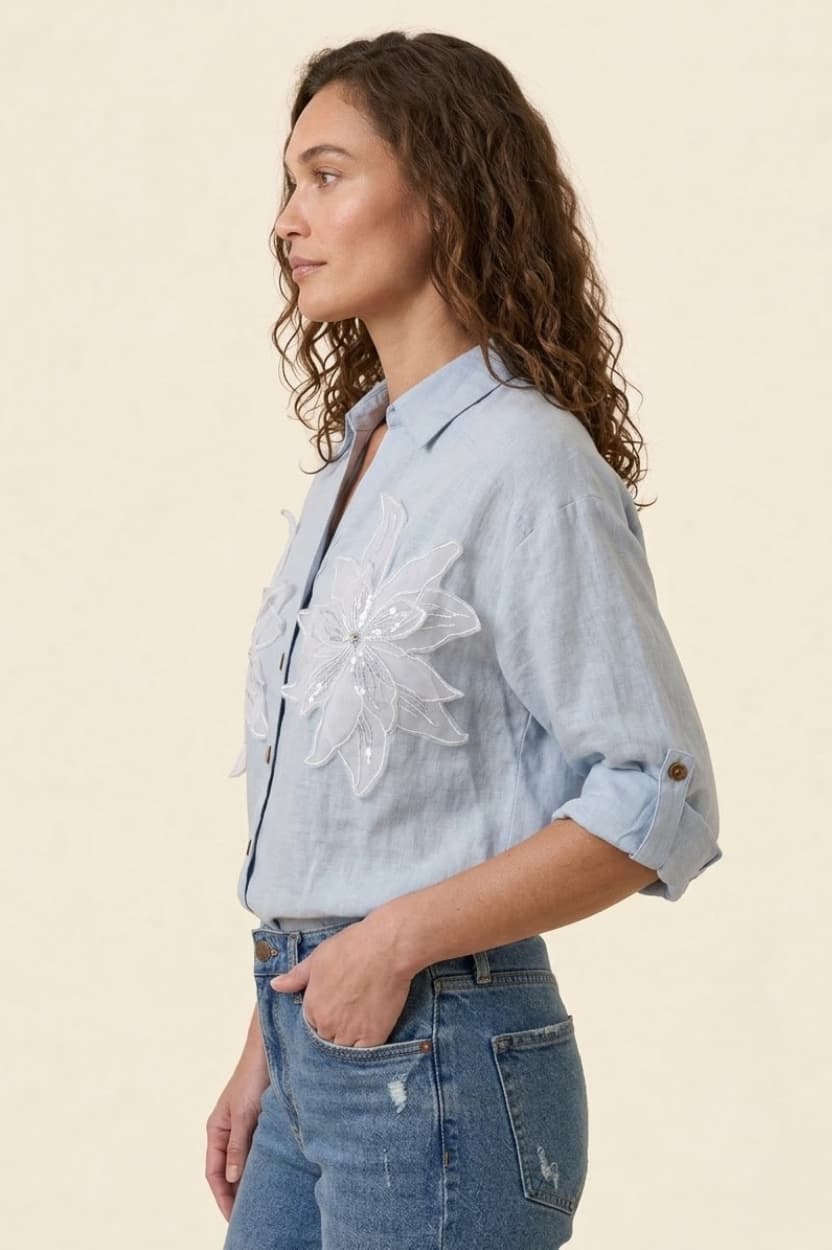 Woman wearing a light blue denim shirt with floral embroidery and rolled-up sleeves, standing against a beige background.