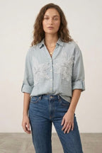 Woman wearing a light blue embroidered shirt and blue jeans against a plain background